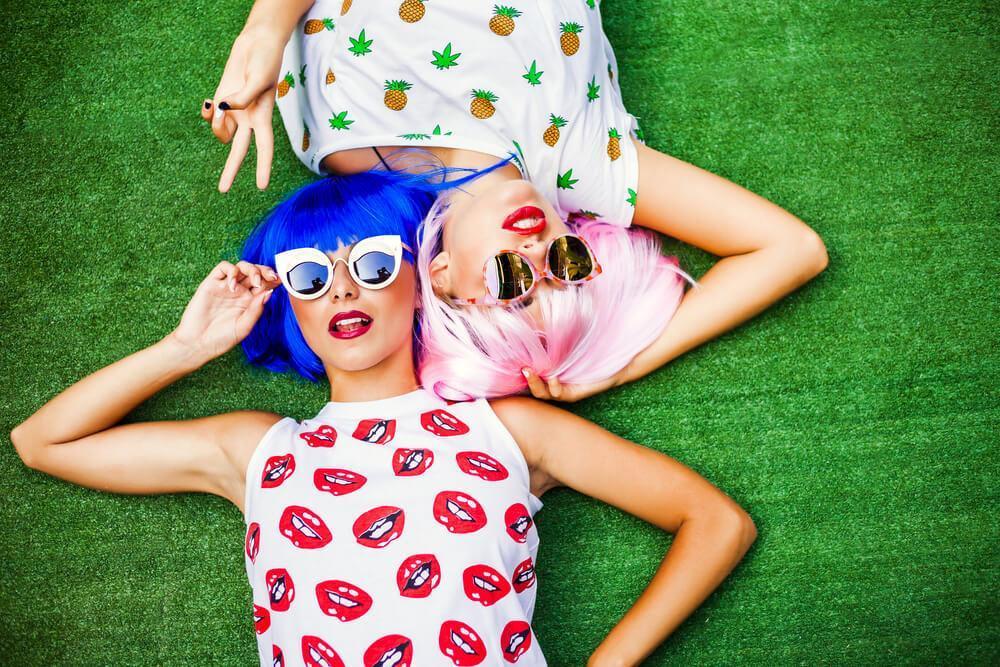 Two young woman with colorful hair wigs and fashionable sunglasses lying on the grass - look good feel good