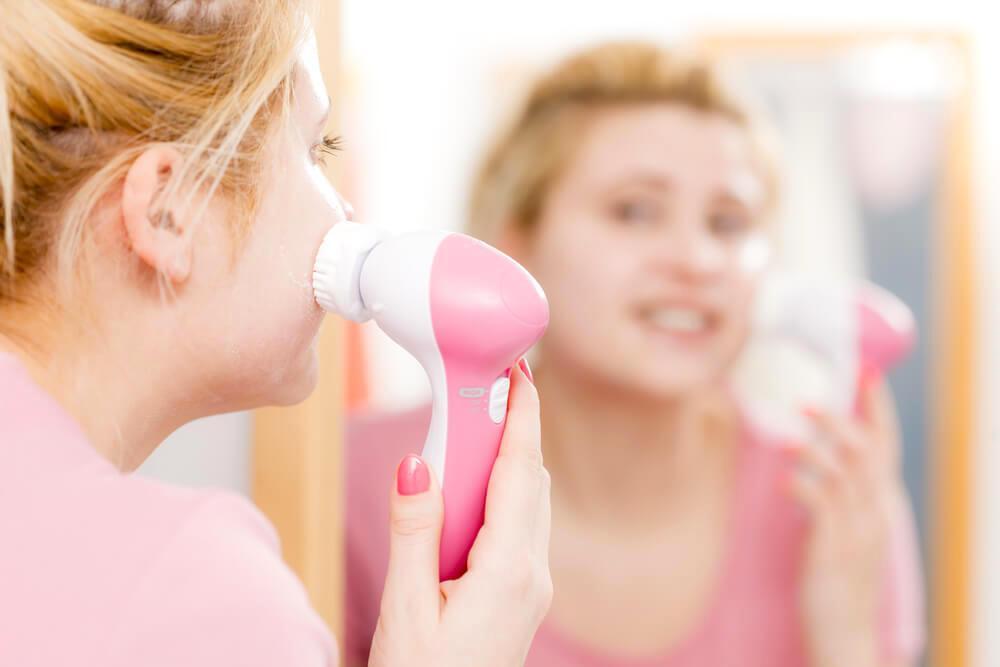 Young woman using facial brush in front of bathroom mirror