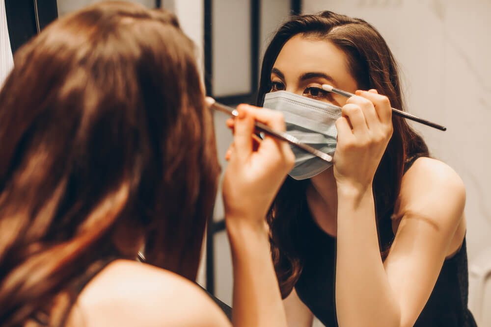Woman applying eye makeup in mirror while wearing face mask